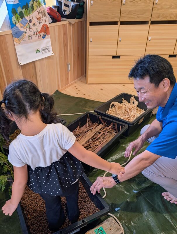 ヒノキの球果の上を歩くことをためらうお子さんをサポートする福ちゃん